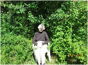 Janet Pattinson, sitting at the edge of our “bush”.  Saskatoon, Mock Orange and Ocean Spray surround Janet and Honeysuckle, Oregon Grape and a Fir tree found behind all planted by Mother Nature