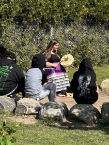 Lisa Ross, local educator and Indigenous Knowledge Holder sitting in a circle with kids in nature, holding a drum