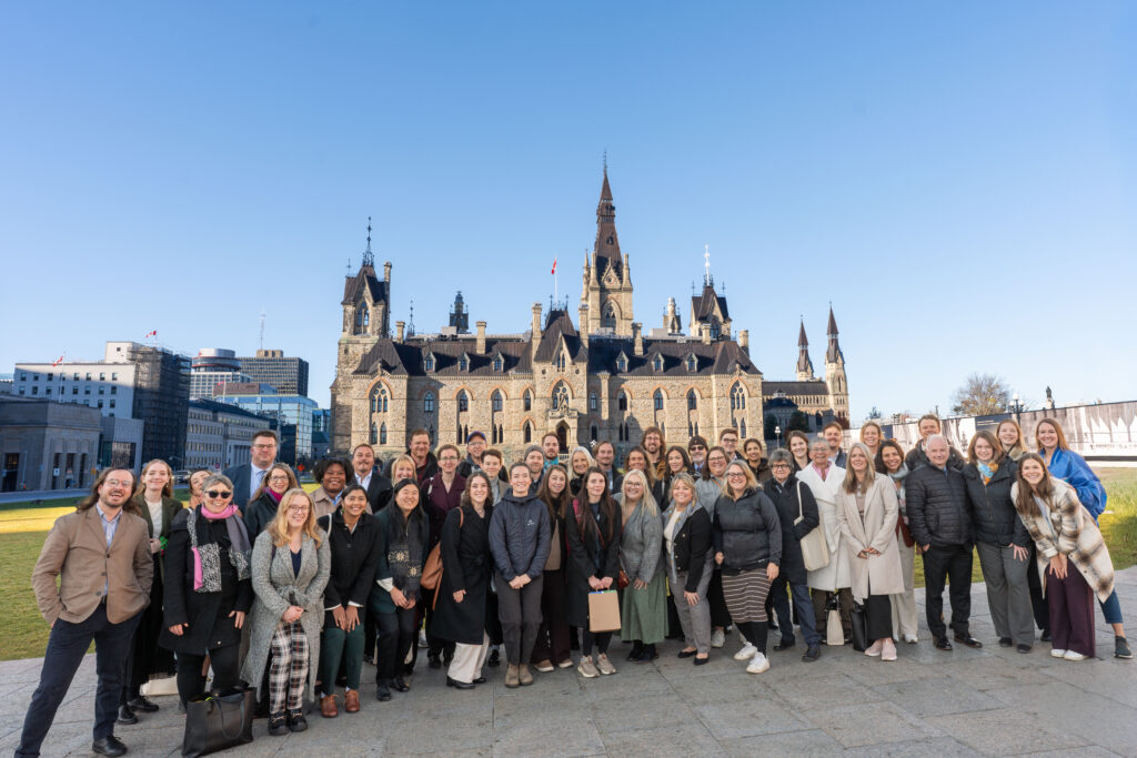 A wide group portrait of several dozen people standing on a stone plaza in front of the historic, Gothic-style Centre Block of Parliament Hill in Ottawa, Canada.The group is diverse in age and gender, dressed in business-casual attire, and smiling toward the camera. In the background, the grand stone architecture of the Parliament building is bathed in bright afternoon sunlight under a clear, pale blue sky. Modern city buildings are visible to the far left, creating a contrast between the historic landmark and the contemporary urban landscape.