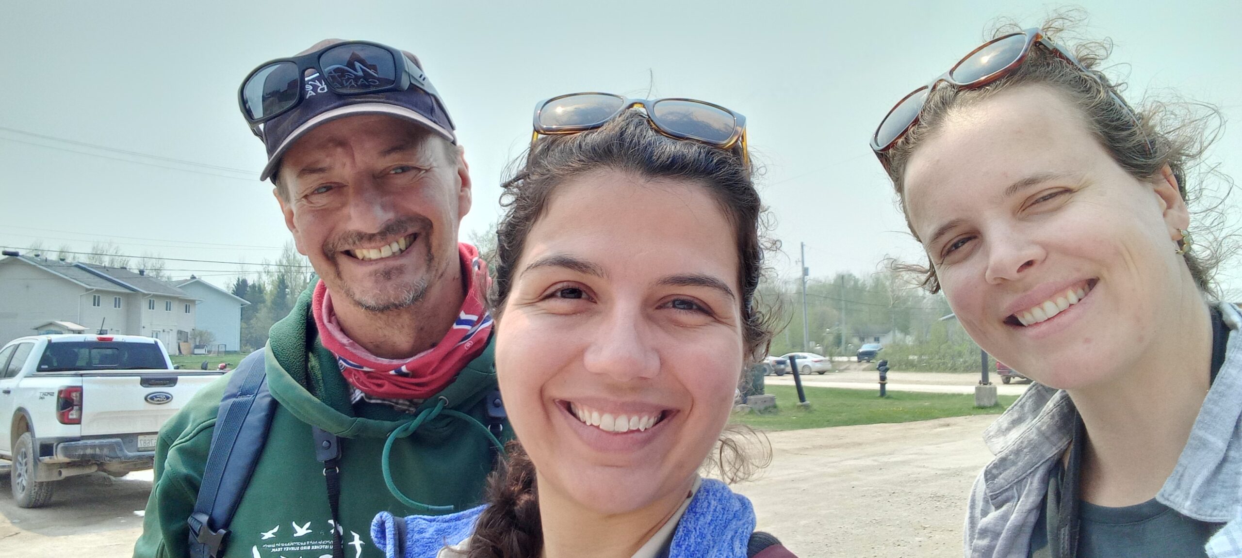 Three people smiling for a selfie outdoors, with two wearing sunglasses and one wearing a cap. A pickup truck and trees are visible in the background. The three people are the Nature Canada Naturalist team. From Left to right; Ted Cheskey, Priscilla Santos, and Lara Falkiner