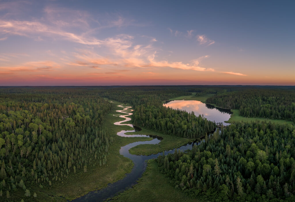 An aerial, wide-angle shot of a winding river snaking through a lush, dense green forest at sunset. The river creates a sharp S-curve in the center of the frame, leading toward a larger, still lake on the right that reflects the soft orange and pink hues of the sky. The horizon is topped with wispy, feathery clouds caught in the glow of the setting sun, fading into a deep twilight blue.
